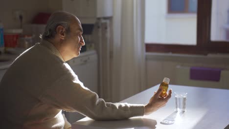 Elderly-sick-man-looking-at-medicine-bottle-in-kitchen-at-home.